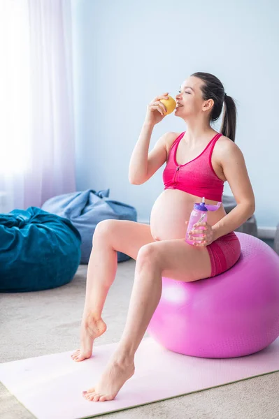 Zwangere vrouw zonder gezicht die een appel eet na yoga. Verwachte moeder bijt fruit en drinkt water na Pilates. Meisje zittend op een fitness bal. — Stockfoto