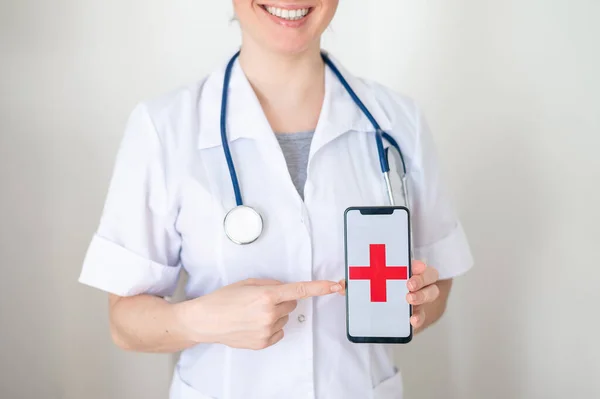 Nerozpoznatelná doktorka drží na obrazovce smartphone s červeným křížem. Sestra se usměje a ukáže na obrazovku mobilního telefonu. — Stock fotografie
