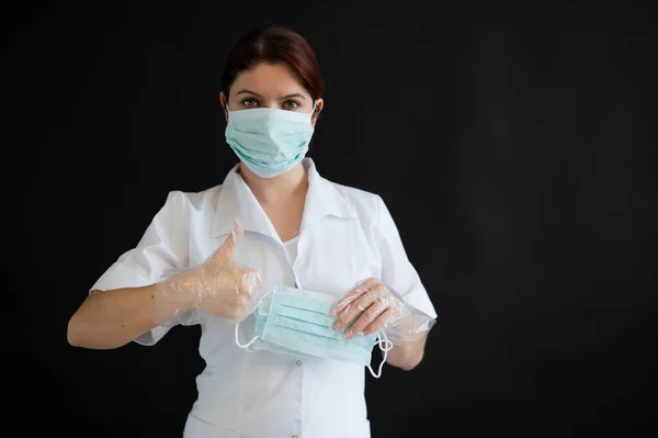 Una joven doctora de uniforme sostiene una máscara médica sobre un fondo negro. Enfermera muestra pulgar ok. Protección contra Coronovirus . — Foto de Stock