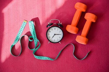 Pembe yoga minderinde iki turuncu tünel mezurası ve çalar saat. Spor yapma ve yazın kilo verme zamanı geldi. Evde ekipmanlı spor.
