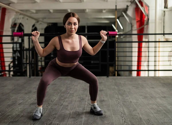 Una hermosa mujer europea se sienta en cuclillas con una barra en los hombros. Chica joven en ropa deportiva haciendo ejercicios en los músculos de las nalgas . —  Fotos de Stock