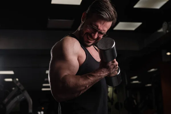 Un hombre con esfuerzo hace un ejercicio para bíceps y tríceps con una mancuerna. Culturista en relieve. Musculoso chico con gafas en el gimnasio . —  Fotos de Stock