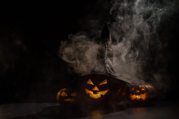 Tres espeluznantes calabazas sonrientes de Halloween brillan en la oscuridad entre la niebla. jack-o-lantern en un sombrero de bruja sobre un fondo negro en humo. — Foto de Stock