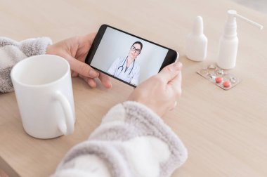 Cep telefonunda bir doktorla çevrimiçi istişare yapan tanınmayan bir kadın. Kız hasta ve evden gelen bir görüntülü aramada uzman doktorla konuşuyor. Ekranı kapat.