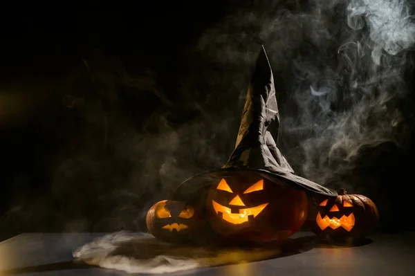 Three creepy Halloween grinning pumpkins glow in the dark among the fog. jack-o-lantern in a witch hat on a black background in smoke.