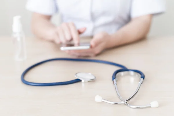 O médico está escrevendo no smartphone durante o intervalo. cardiologista feminino dá consulta on-line por telefone celular. Estetoscópio e mão anti-séptico na área de trabalho . — Fotografia de Stock