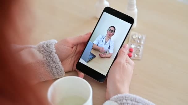 El médico jura al paciente para una consulta en línea por teléfono. La chica está enferma y habla con el médico de cabecera en una videollamada desde casa . — Vídeo de stock