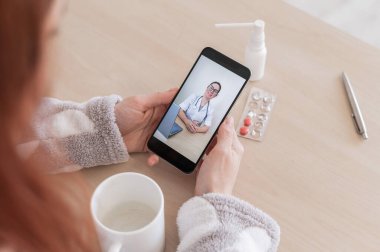 Cep telefonunda bir doktorla çevrimiçi istişare yapan tanınmayan bir kadın. Kız hasta ve evden gelen bir görüntülü aramada uzman doktorla konuşuyor. Ekranı kapat.