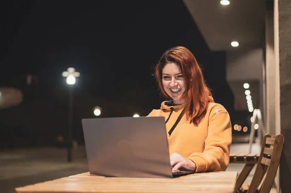 Egy nő távirányítással dolgozik egy laptopnál egy nyári kávézóban késő este. Happy Girl tanul, miközben egy üres utcán ül egy fa asztalnál. Női szabadúszó pulóverben esténként — Stock Fotó