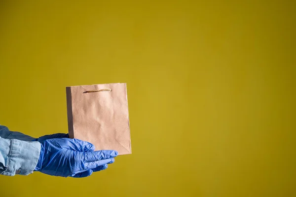 Closeup of female hands in gloves and a denim shirt. Delivery man holds an empty paper bag on a yellow background. Craft packaging for takeaway snack. Antimicrobial protection. — Stock Photo, Image