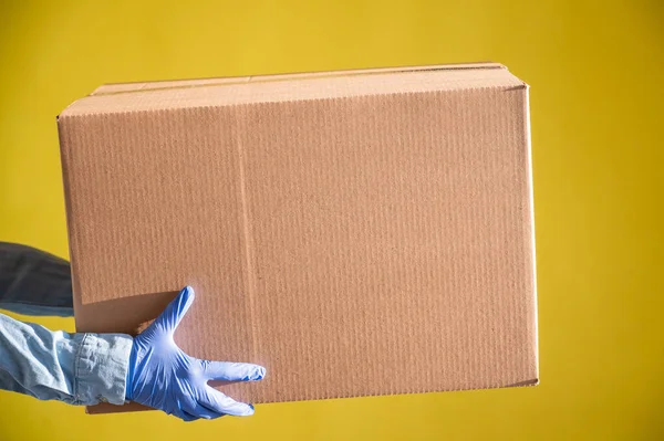 Closeup of female hands in gloves and a denim shirt. The delivery man passes a cardboard box to the customer on a yellow background. Antimicrobial protection in quarantine. Cropped. — Stock Photo, Image