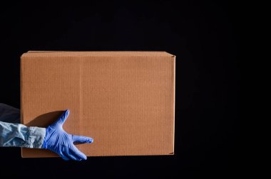 Eldivenli ve kot gömlekli kadın elleri. Teslimatçı karton kutuyu siyah arka planda müşteriye verir. Karantinada antimikrop koruması. Kırpılmış.