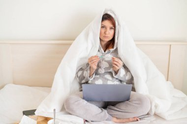 Yataktaki kadın kafasıyla battaniyeyle örtülmüş ve sıcaklığı ölçüyor. Soğuk algınlığı olan bir kız dizlerinin üzerinde dizüstü bilgisayarla yatak odasında oturuyor..