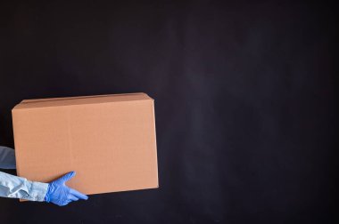 Eldivenli ve kot gömlekli kadın elleri. Teslimatçı karton kutuyu siyah arka planda müşteriye verir. Karantinada antimikrop koruması. Kırpılmış.