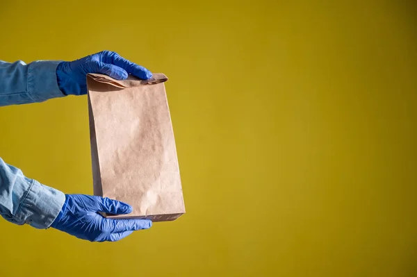 Closeup of female hands in gloves and a denim shirt. Delivery man holds an empty paper bag on a yellow background. Craft packaging for takeaway snack. Antimicrobial protection. — Stock Photo, Image