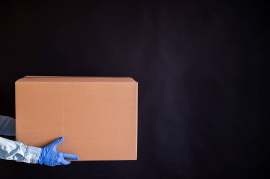 Eldivenli ve kot gömlekli kadın elleri. Teslimatçı, müşteriye siyah arka planda bir karton kutu tutuyor. Bir salgında mikrop önleyici koruma.