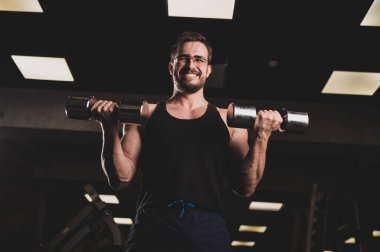 Çabalayan bir adam, pazılar ve trisepsler için halterli egzersiz yapar. Kabartmalı vücut geliştirici gülümsemesi. Spor salonunda gözlüklü kaslı adam..