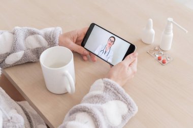 Cep telefonunda bir doktorla çevrimiçi istişare yapan tanınmayan bir kadın. Kız hasta ve evden gelen bir görüntülü aramada uzman doktorla konuşuyor. Ekranı kapat.