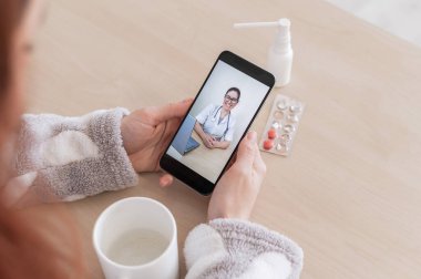 Cep telefonunda bir doktorla çevrimiçi istişare yapan tanınmayan bir kadın. Kız hasta ve evden gelen bir görüntülü aramada uzman doktorla konuşuyor. Ekranı kapat.