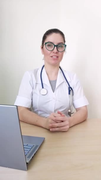 Friendly doctor gestures and talks to the camera. Portrait of a treating doctor sitting at a desk and giving online advice on disease prevention. — Stock Video