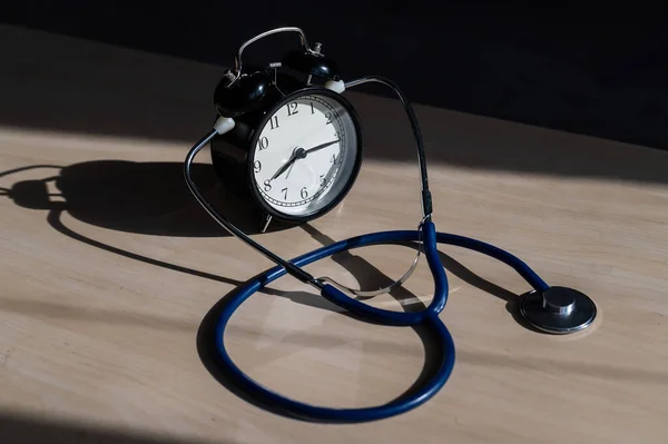 Relógio de alarme mecânico preto velho clássico com estetoscópio em uma mesa de madeira. Está na hora de verificar o coração. Relógio de mesa e um dispositivo para medir a pressão arterial . — Fotografia de Stock