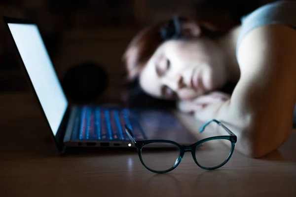 Trött kvinna sover på arbetsplatsen nära laptop. Flickan slumrade till för fjärrarbete. Överarbetad elev somnade på bordet. Glasögon i förgrunden. — Stockfoto