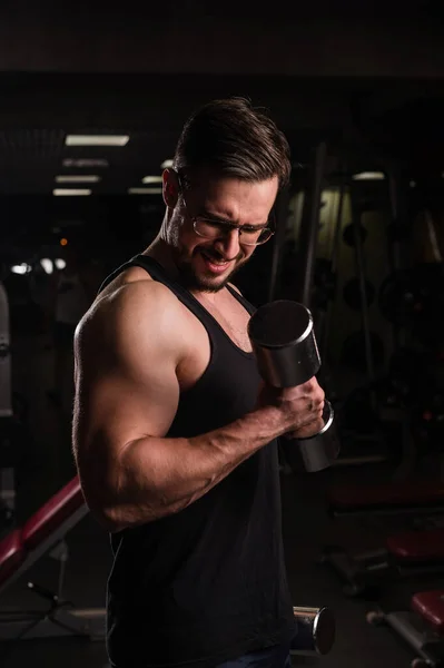 Un hombre con esfuerzo hace un ejercicio para bíceps y tríceps con una mancuerna. Culturista en relieve. Musculoso chico con gafas en el gimnasio . —  Fotos de Stock