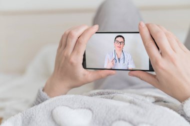 Bir kadın evde karantinada ve bir virologla video konferansı yapıyor. Hasta elinde bir telefonla yatakta uzanıyordu. Doktordan telefon var..