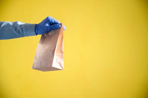 Closeup of female hands in gloves and a denim shirt. Delivery man passes an empty paper bag on a yellow background. Craft packaging for takeaway snack. Antimicrobial protection. — Stock Photo, Image
