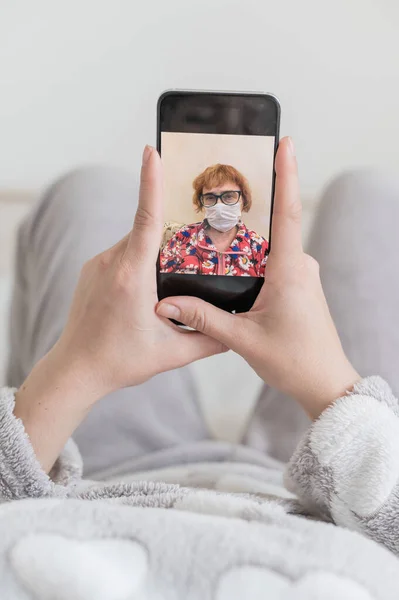 The girl during quarantine lies in bed and makes a video call on the phone to her mother. Close-up of female hands holding a smartphone with an elderly woman in a medical mask on the screen.