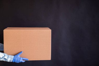 Eldivenli ve kot gömlekli kadın elleri. Teslimatçı, müşteriye siyah arka planda bir karton kutu tutuyor. Bir salgında mikrop önleyici koruma.