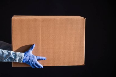 Eldivenli ve kot gömlekli kadın elleri. Teslimatçı karton kutuyu siyah arka planda müşteriye verir. Karantinada antimikrop koruması. Kırpılmış.