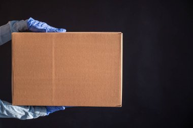 Eldivenli ve kot gömlekli kadın elleri. Teslimatçı, müşteriye siyah arka planda bir karton kutu tutuyor. Bir salgında mikrop önleyici koruma.