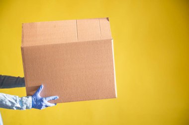 Eldivenli ve kot gömlekli kadın elleri. Teslimatçı müşteriye sarı arka planda bir karton kutu verir. Karantinada antimikrop koruması. Kırpılmış.