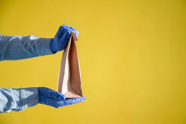 Closeup of female hands in gloves and a denim shirt. Delivery man holds an empty paper bag on a yellow background. Craft packaging for takeaway snack. — Stock Photo, Image