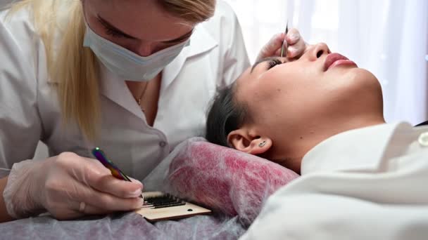 Asian woman lies on a couch during eyelash extension procedure. Master and model on a beauty procedure. — Stock Video