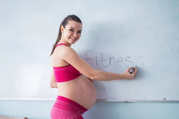 Seorang wanita hamil dalam pakaian olahraga menulis kata kebugaran pada papan penanda putih. Seorang gadis di trimester 3 masuk untuk olahraga dengan perut kosong. Kursus untuk ibu hamil. — Stok Foto