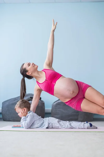 A pregnant woman in sportswear does gymnastics while her son plays in the mobile. Girl on yoga with her child. Fitness mom and baby. — Stock Photo, Image