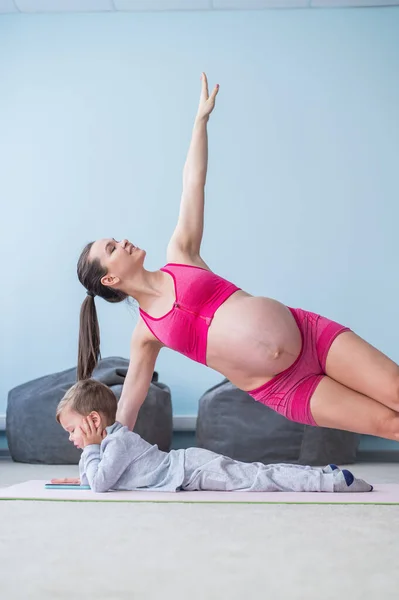 Una donna incinta in abbigliamento sportivo fa ginnastica mentre suo figlio gioca nel cellulare. Ragazza sullo yoga con il suo bambino. Fitness mamma e bambino. — Foto Stock