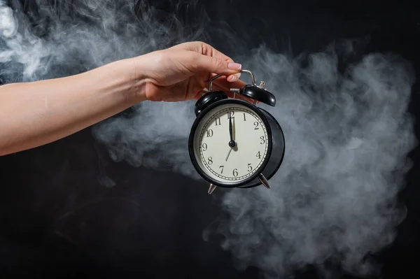 Primer plano de una mano femenina sosteniendo un reloj sobre un fondo negro en humo. Reloj despertador a medianoche en una niebla mística . —  Fotos de Stock