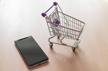 Alışveriş arabasının üst görüntüsü ve ahşap bir masada cep telefonu. Akıllı telefon çevrimiçi alışveriş. İnternetten alışveriş.