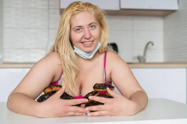 Een dikke blonde met grote borsten eet een donut met chocolade in de keuken. Een zwaarlijvige jonge vrouw heeft twee borden vol zoete gebak. Meisje met een medisch masker. — Stockfoto