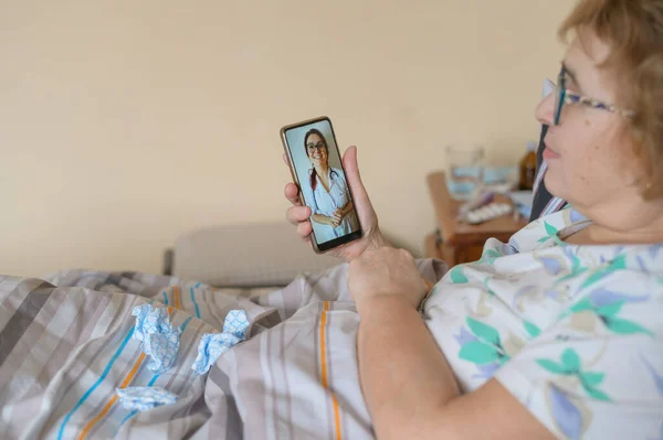 An elderly woman is sick with coronavirus and is in quarantine. A pensioner lies in bed and consults a doctor online about a viral disease.