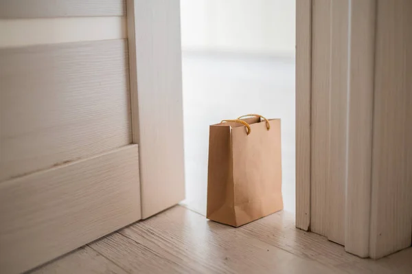 The concept of contactless delivery during the quarantine period. Kraft paper bag with online purchase at the open entrance door in case of danger of the spread of coronavirus. — Stock Photo, Image