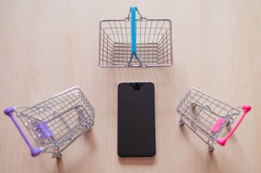 Bir cep telefonunun üst görüntüsü iki yemek arabası ve ahşap bir masada bir sepet. İnternetten akıllı telefondan alışveriş. Küçük Resimler.