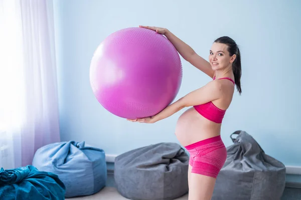 Een zwangere vrouw in sportkleding doet gymnastiek voor zwangere vrouwen. Het meisje is bezig met fitness met een bal voor aanstaande moeders. Laatste trimester. — Stockfoto