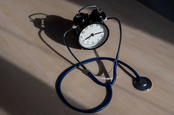 Relógio de alarme mecânico preto velho clássico com estetoscópio em uma mesa de madeira. Está na hora de verificar o coração. Relógio de mesa e um dispositivo para medir a pressão arterial . — Fotografia de Stock