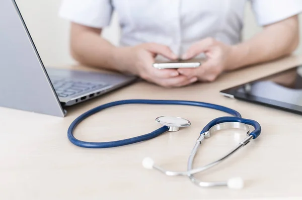 Felismerhetetlen női orvos ül egy asztalnál, és okostelefont használ. Egy gyakorló kezének közelsége egy telefonnal. Stethoscope tabletta és laptop az asztalon. — Stock Fotó