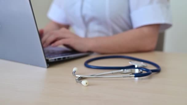 Kobieta w płaszczu medycznym na biurku, pisząca na laptopie. Pielęgniarka wypełnia elektroniczną kartę pacjenta. — Wideo stockowe