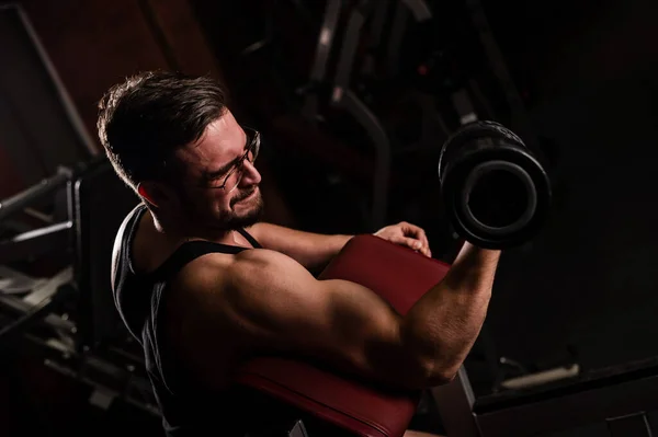 Um homem bonito com óculos a fazer exercício para bíceps com uma campainha. O tipo está envolvido em musculação. Treinador no ginásio com braços musculares . — Fotografia de Stock